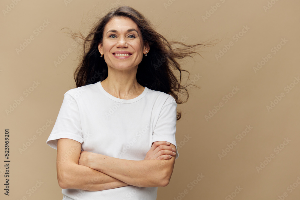 Obraz premium beautiful, woman with long, beautiful, well-groomed hair that develops in the wind, stands smiling sweetly at the camera, with her arms folded across her chest. Horizontal photo on a beige background.