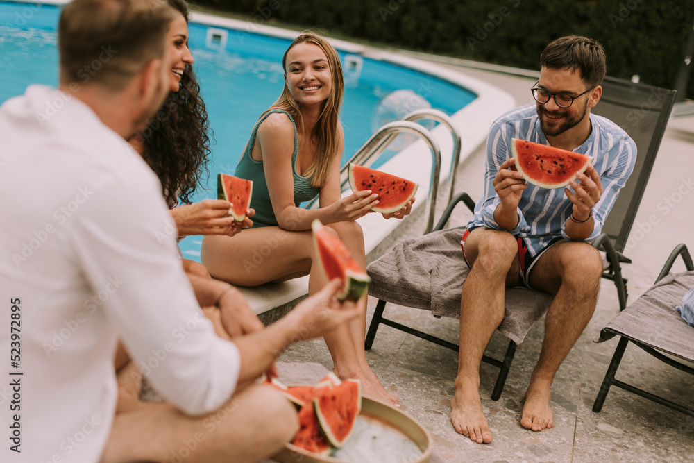 Young people sitting by the swimming pool and eating watermelon in the house backyard