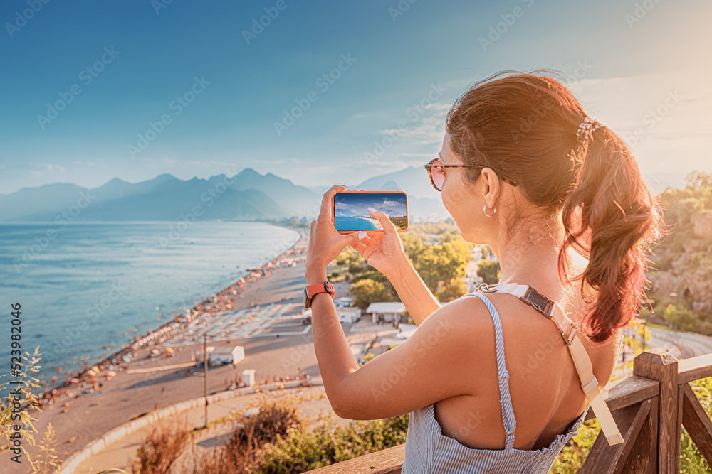 Fototapeta premium Travel blogger girl takes high-quality and vivid photos on the camera of his new expensive smartphone of the famous Konyaalti beach from a scenic viewpoint in Antalya, Turkey