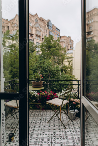 a coffee table and two chairs on an open balcony with a city view. blooming flowers in pots decorate the balcony 