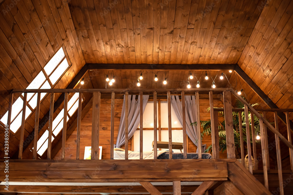 Scandinavian style interior at home. Country wooden house is spacious on two levels. Room with wooden stairway leading to second floor of modern flat. Bedroom with lamp garlands on second floor. Scandinavian style interior at home. Country wooden house is spacious on two levels. Room with wooden stairway leading to second floor of modern flat. Bedroom with lamp garlands on second floor.