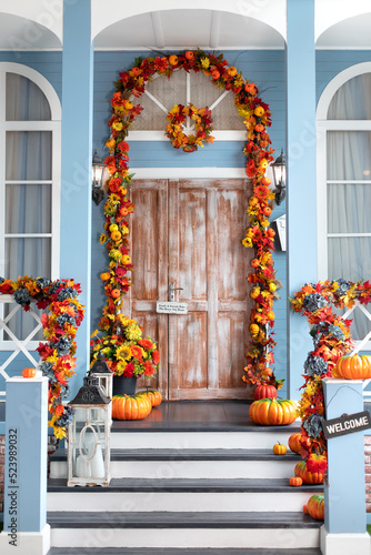 House entrance staircase decorated for autumn holidays, fall flowers and pumpkins. Cozy wooden porch of the house with pumpkins in fall time. Halloween design home with yellow fall leaves and lanterns