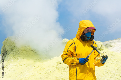 woman volcanologist on the background of a smoking fumarole examines a sample of a sulfur mineral with a geological hammer
