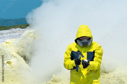 man volcanologist on the background of a smoking fumarole examines a sample of a sulfur mineral with a geological hammer