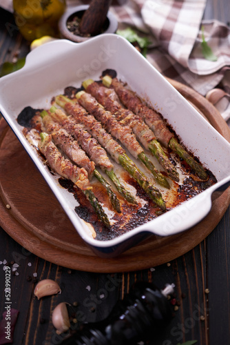 ceramic baking dish with baked aspargus covered with bacon on wooden kitchen table