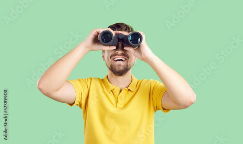 Happy joyful young man in casual yellow T shirt standing isolated on green background, holding modern binoculars, looking ahead and smiling. Making discoveries and having fun while travelling concept