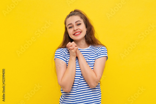 Portrait of cute young brunette woman in striped t shirt, smiling and holding hands under chin, has happy expression, gazing camera thankful. Indoor studio shot on yellow background