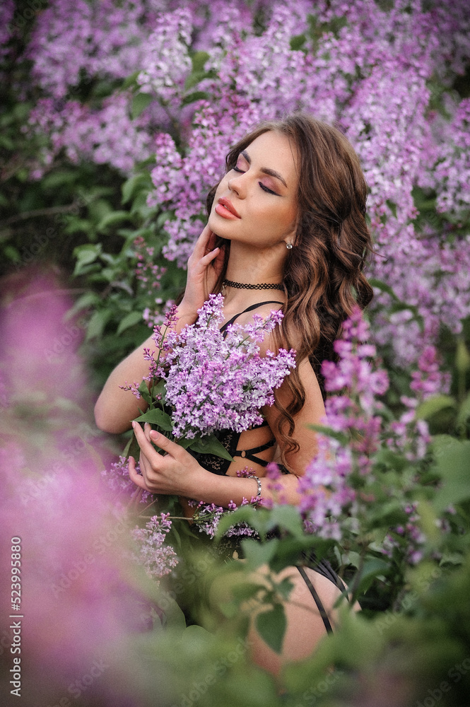 a beautiful girl with makeup and hair styling in underwear stands in a garden with lilacs on a summer evening a beautiful girl with makeup and hair styling in underwear stands in a garden with lilacs on a summer evening