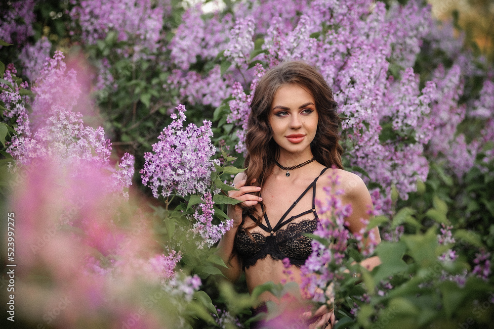 a beautiful girl with makeup and hair styling in underwear stands in a garden with lilacs on a summer evening a beautiful girl with makeup and hair styling in underwear stands in a garden with lilacs on a summer evening