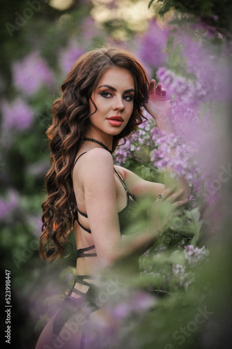 Fototapeta Naklejka Na Ścianę i Meble -  a beautiful girl with makeup and hair styling in underwear stands in a garden with lilacs on a summer evening