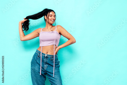 Young beautiful smiling female in trendy summer jeans and top clothes. Carefree woman posing near blue wall in studio. Sexy positive model having fun indoors. Cheerful and happy. Holding her hair tail