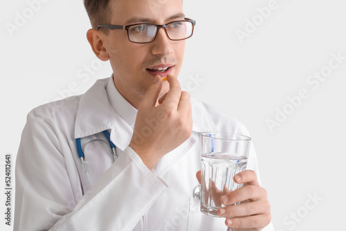 Young doctor with glass of water taking pill on white background