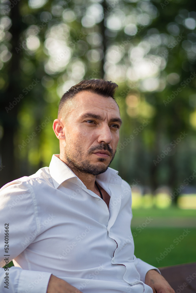 A pensive handsome man in a white shirt is resting sitting on a bench. Young man in city park, warm sunny summer day. Warm soft light, close-up. A pensive handsome man in a white shirt is resting sitting on a bench. Young man in city park, warm sunny summer day. Warm soft light, close-up.