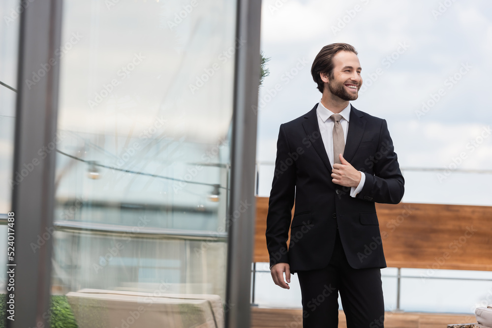 Young groom smiling while adjusting jacket on terrace.