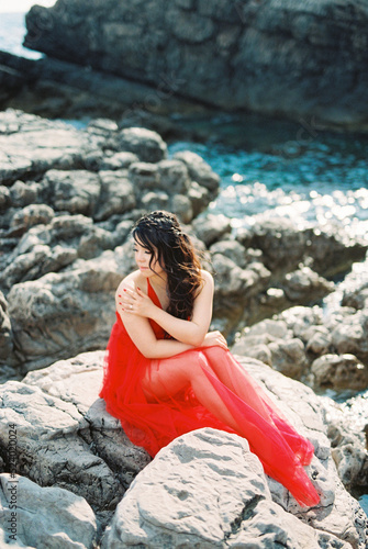 Young woman in a red dress sits on the rocks near the water and looks away © Nadtochiy