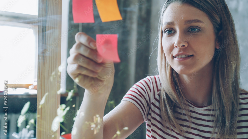 Close-up of pretty young woman with blond hair sticking colored memos on glass board in modern office. She is looking at papers and smiling. Creative work concept. Close-up of pretty young woman with blond hair sticking colored memos on glass board in modern office. She is looking at papers and smiling. Creative work concept.
