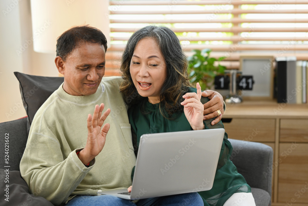 Happy aged spouses making video call with family or friends on laptop while spending time together at home Happy aged spouses making video call with family or friends on laptop while spending time together at home
