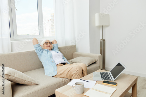 a carefree, stylish, happy elderly woman in stylish home clothes is sitting on the couch relaxing while working on a laptop at home