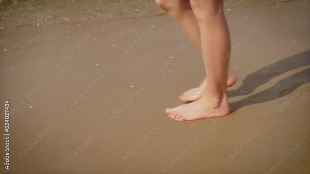 Slim female legs and feet walking along sea water waves on sandy beach. Pretty woman walks at seaside surf. Splashes of water and foam. Freedom lifestyle and traveling in holiday concept Slim female legs and feet walking along sea water waves on sandy beach. Pretty woman walks at seaside surf. Splashes of water and foam. Freedom lifestyle and traveling in holiday concept