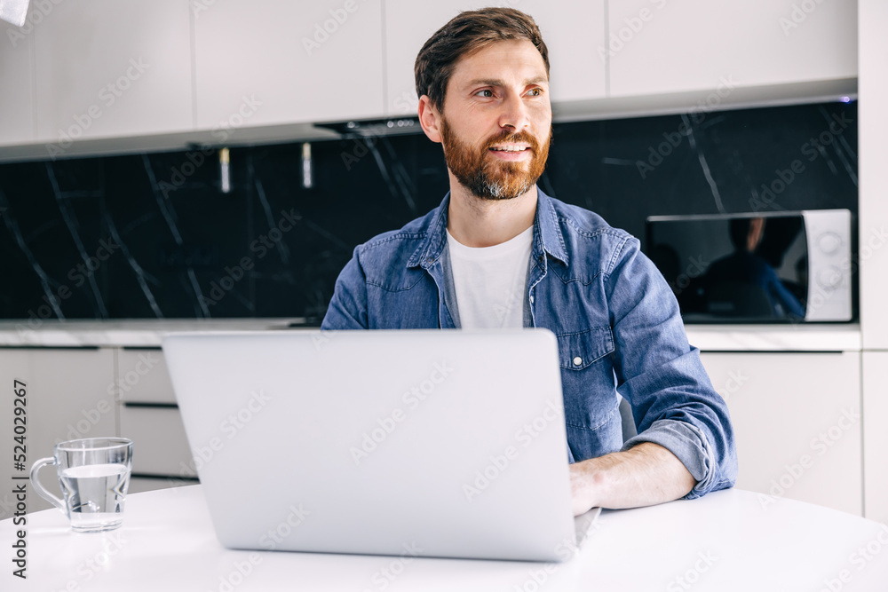 Smiling professional businessman using laptop computer working from home office. Happy man sitting at the kitchen table looking to the side Smiling professional businessman using laptop computer working from home office. Happy man sitting at the kitchen table looking to the side