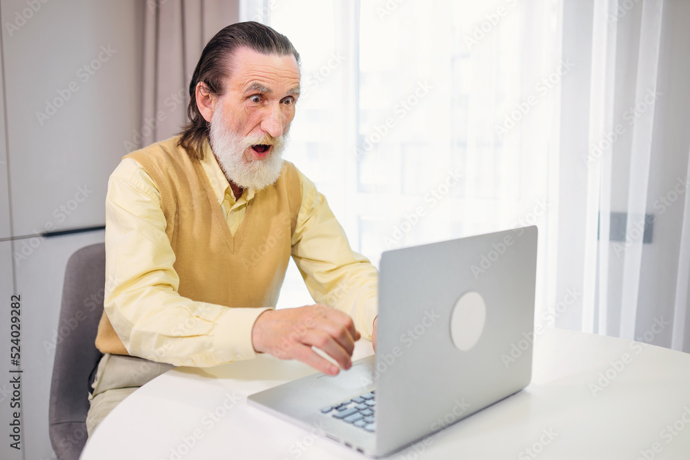 A shocked, stressed elderly man looks at a laptop, reads incredible online news at home, an old grandfather feels unpleasantly surprised by an unexpected problem A shocked, stressed elderly man looks at a laptop, reads incredible online news at home, an old grandfather feels unpleasantly surprised by an unexpected problem