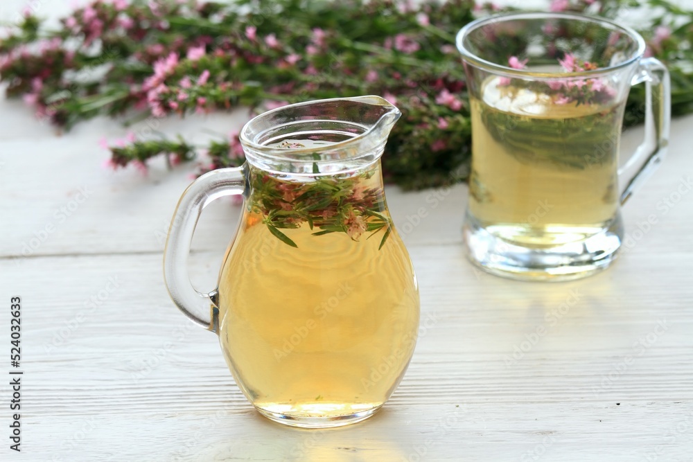 Herbal tea in glass with fresh flowers of hyssopus, lat. Hyssopus officinalis. Homemade tea from hyssopus and hot water in a jug and glass on white wooden table. Herbal tea in glass with fresh flowers of hyssopus, lat. Hyssopus officinalis. Homemade tea from hyssopus and hot water in a jug and glass on white wooden table.