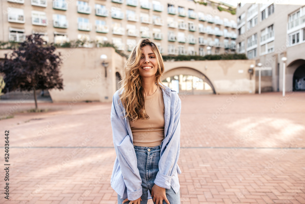 Stylish young caucasian girl smiling looking at camera spending leisure time outdoors. Blonde woman with wavy hair wears casual clothes. Sincere emotions lifestyle concept