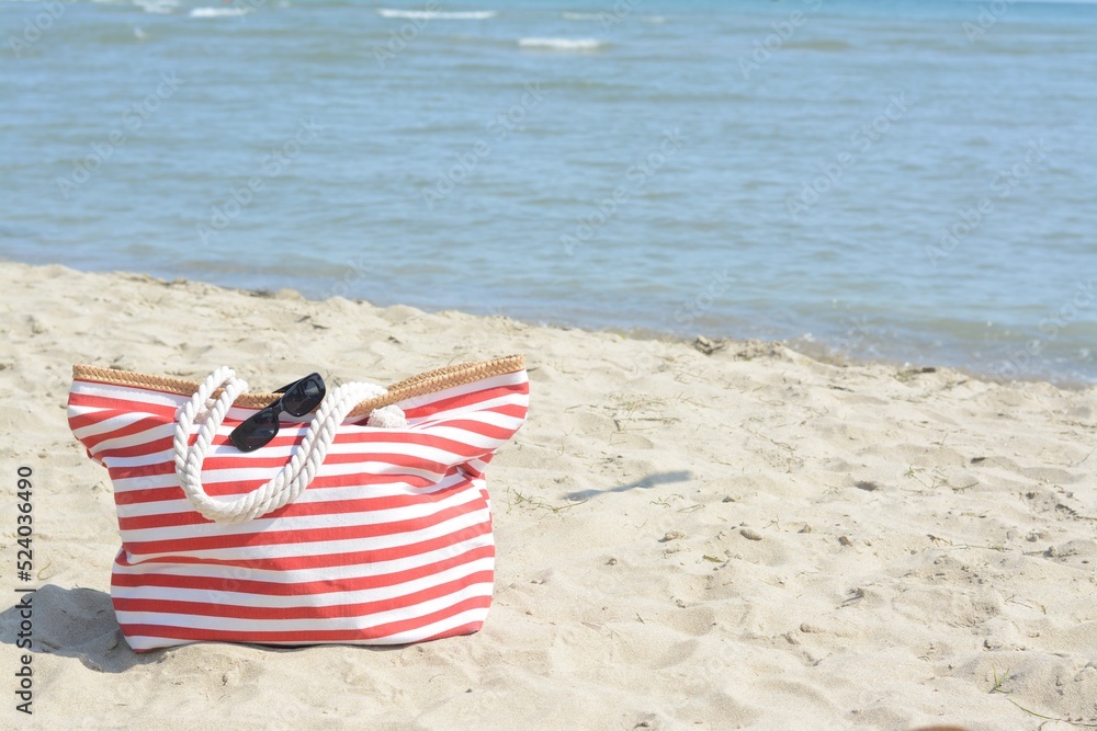 Stylish striped bag with sunglasses on sandy beach near sea, space for text Stylish striped bag with sunglasses on sandy beach near sea, space for text
