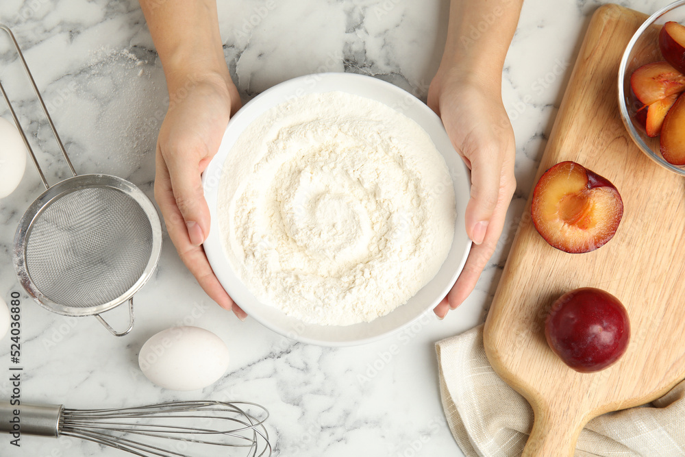 Fototapeta premium Woman holding bowl with flour at white marble table, top view. Cooking of delicious plum cake