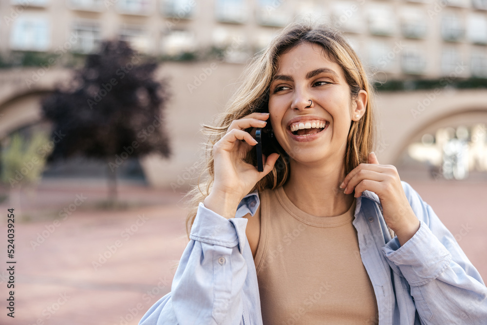 Positive young caucasian girl in good mood is talking on phone standing on street. Blonde smiles with her teeth, wears shirt. Lifestyle concept, use Positive young caucasian girl in good mood is talking on phone standing on street. Blonde smiles with her teeth, wears shirt. Lifestyle concept, use
