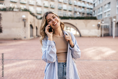 Cheerful young caucasian woman talking on phone spending time outdoors during day. Blonde smiles, wears casual clothes. Technology concept