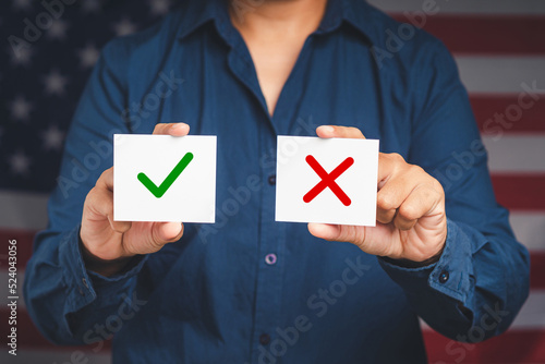 Close-up of hands holding two white papers with a green checkmark and red cross while standing on the American flag background
