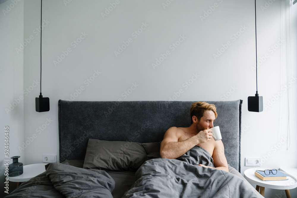 Fototapeta premium Young bearded shirtless man drinking coffee while lying in bed at home