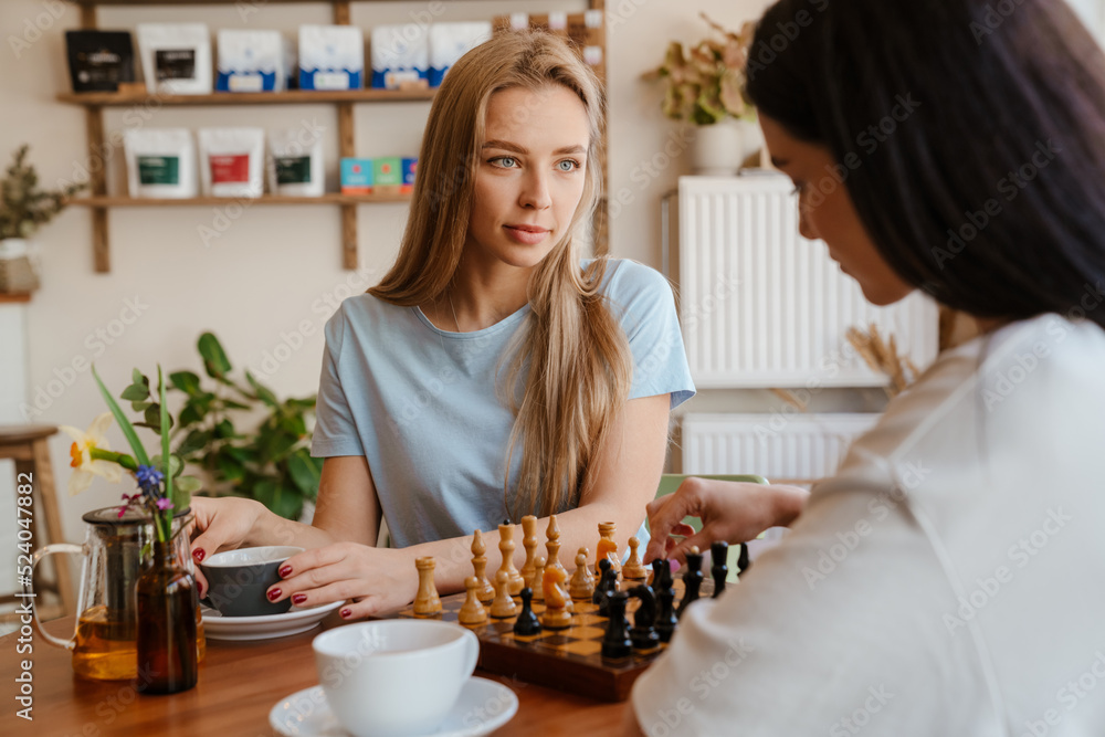 White young women playing chess while drinking tea in cafe