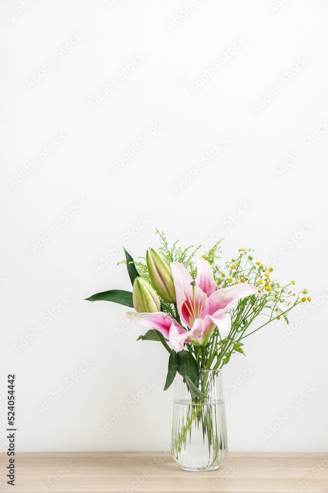 A bouquet of beautiful flowers in a glass vase, with a blossoming pink lily (Lilium), two unopened lily bulbs, and small daisies - displayed on right side of wooden surface against white background