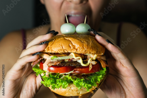 latina girl about to bite a hamburger she is holding with both hands. hamburger with two quail eggs on top imitating eyes. street food on black background. detailed view of ingredients.