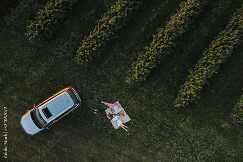 Aerial top-down shot of couple enjoying picnic on road trip in the beautiful fields, man and woman lying on a blanket near the SUV car at sunset