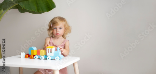 Pretty caucasian 1,2 year old with blond curly hair playing with colourful construction cubes, toys making train, looking at camera.Happy smiling child having fun,early development concept.Copy space.
