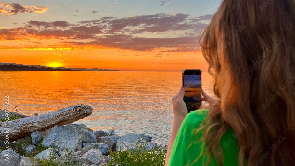 a teenage girl in a Green T-shirt stands with her back to the camera and shoots a beautiful sunset on Pacific Ocean she holds an iPhone 13 phone in her hands blond red hair stones setting sun sea sky a teenage girl in a Green T-shirt stands with her back to the camera and shoots a beautiful sunset on Pacific Ocean she holds an iPhone 13 phone in her hands blond red hair stones setting sun sea sky