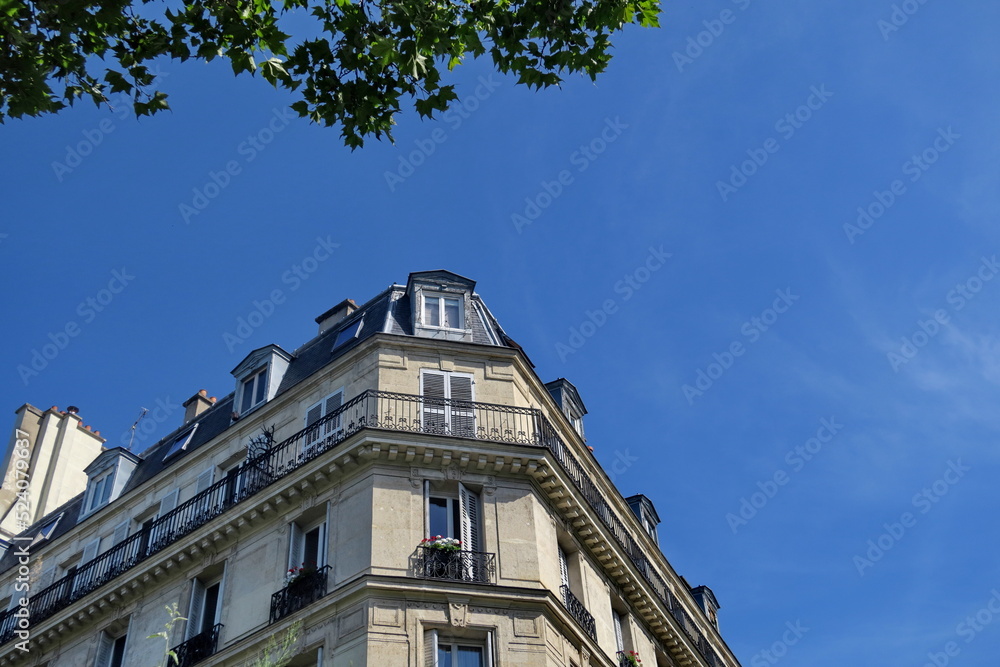 Obraz premium Immeuble ancien en coin de rue. Façade blanche, ciel bleu et feuillage vert.