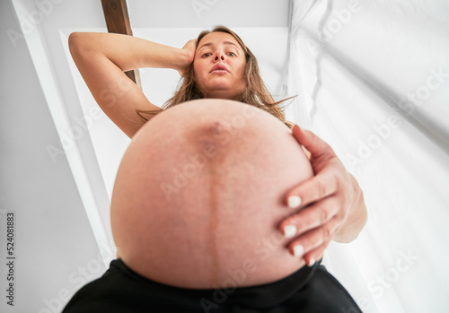 Bottom view of pregnant lady who surprised by size of her belly. Woman putting one hand on her pregnant belly and the other on her head and looking down in dismay.