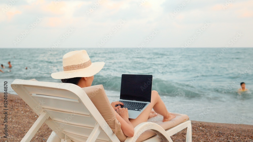 Rear view of freelancer woman working on laptop near the sea. Remote work concept. work on vacation. 4 k