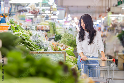 Organic food market. Middle age asian housewife pushing shopping cart walking near vegetables stalls and other agriculture products on marketplace sale. Wife thinking what to buy prepare for dinner. 