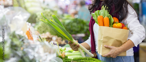 Shopping or Food delivery service concept - Panoramic shot of Woman holding eco friendly reusable shopping bag filled with vegetables and choosing to buy vegetables, Blurred supermarket in background.
