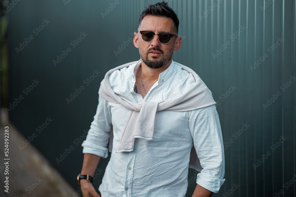 Fashion model handsome young man with trendy hairstyle in sunglasses in white shirt in light jeans posing near vintage wall on street on sunny day. Attractive american guy on walk. Copy space. Fashion model handsome young man with trendy hairstyle in sunglasses in white shirt in light jeans posing near vintage wall on street on sunny day. Attractive american guy on walk. Copy space.