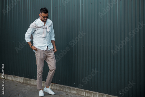 Fashion model handsome young man with trendy hairstyle in sunglasses in white shirt in light jeans posing near vintage wall on street on sunny day. Attractive american guy on walk. Copy space.