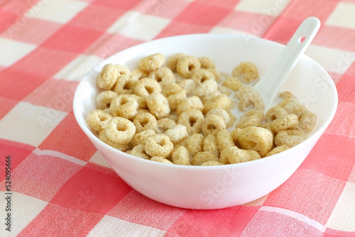 Healthy morning oat cereal. Cheerios in bowl of milk with spoon. Low sugar and high fibre breakfast. Isolated on red and white square patterned tablecloth.