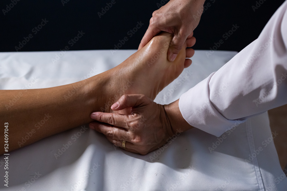 Fototapeta premium Detalhe das mãos de massagista aplicando massagem terapêutica no pé de um paciente que está deitado em uma maca com lençol branco.