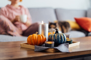 Autumn, fall cozy mood composition for hygge home decor. Small pumpkins, burning candles on tray with gray napkin on the coffee table with resting woman with cat in the living room. Selective focus
