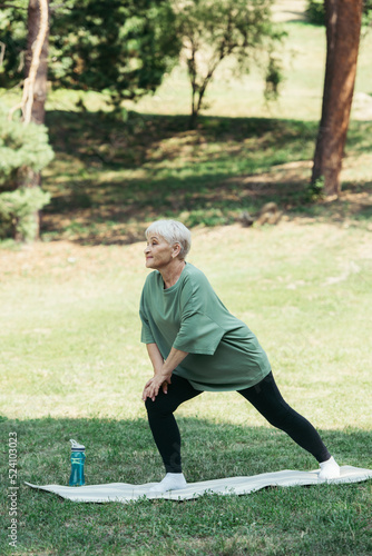 full length of senior woman with grey hair smiling and doing lunges on fitness mat in park.